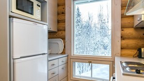 A compact kitchen with a Bosch refrigerator, a microwave, and a stove. There are wooden walls and a window with a snowy view. - Hallantähti mökki by Interhome (Hyrynsalmi)