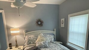 Quarto com cama, mesa de cabeceira, luminária, ventilador de teto, janela com persianas e um objeto decorativo na parede.