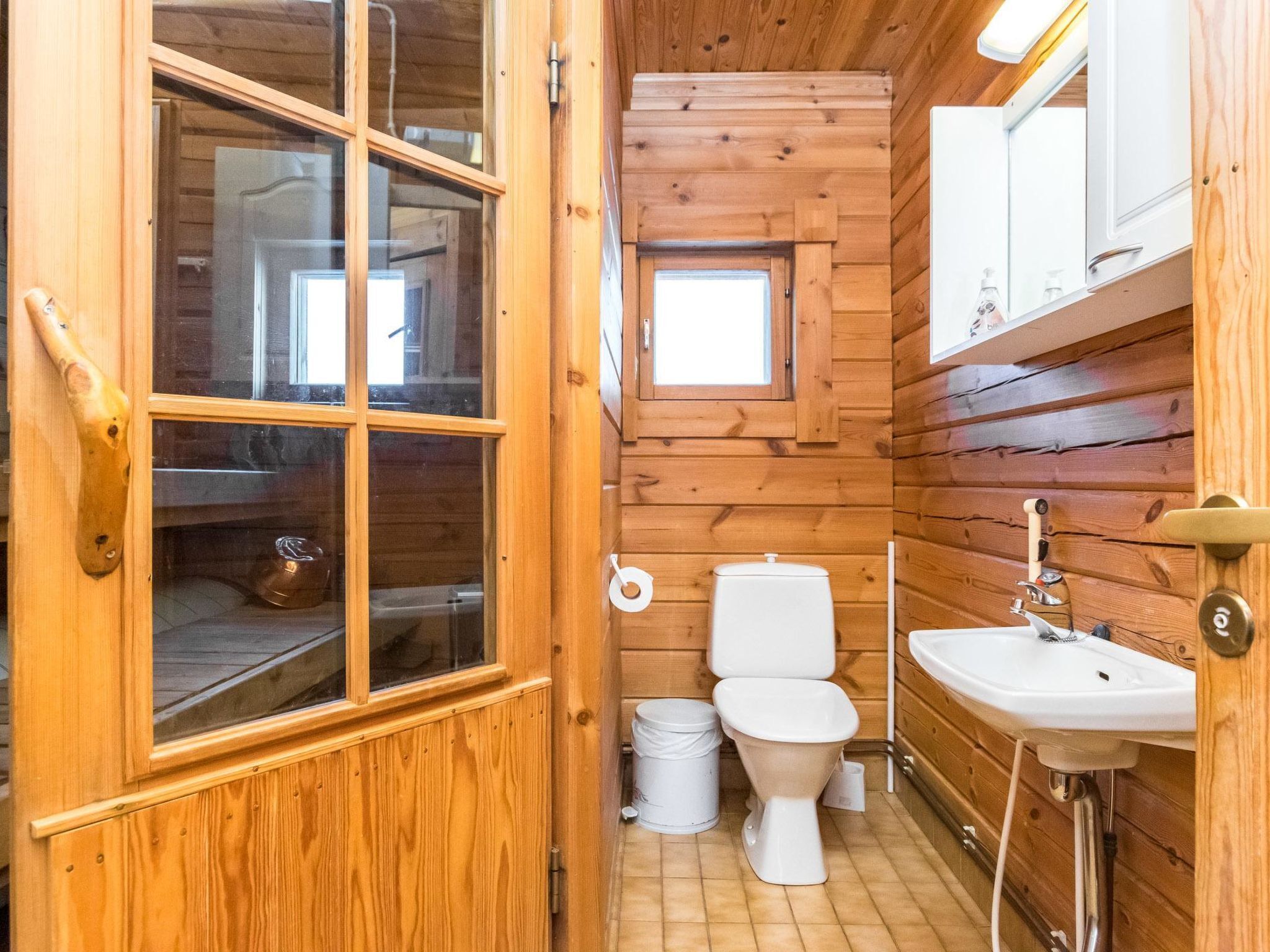 Ein kleines, hölzernes Badezimmer in einer Blockhütte mit Toilette, Waschbecken und einem kleinen Fenster.