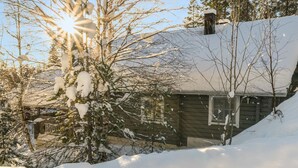 Eine Holzhütte mit schneebedecktem Dach und Schornstein, umgeben von kahlen Bäumen und verschneitem Gelände.