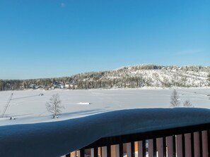 A snow-covered landscape with a clear sky, a wooden deck, and a view of a hill with buildings and trees.