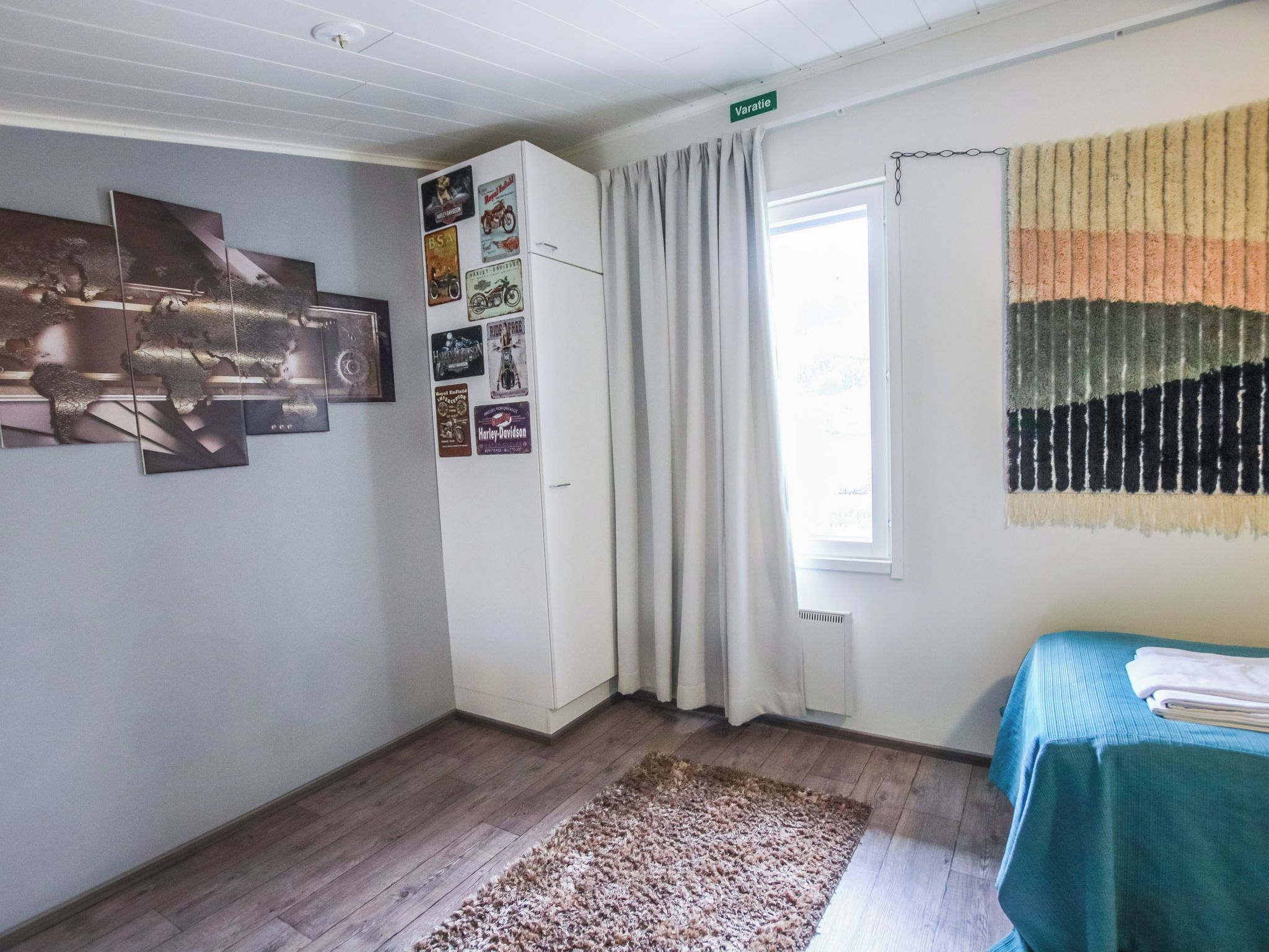 A room with a bed, a window with curtains, a wall with various posters, and a rug on the floor.