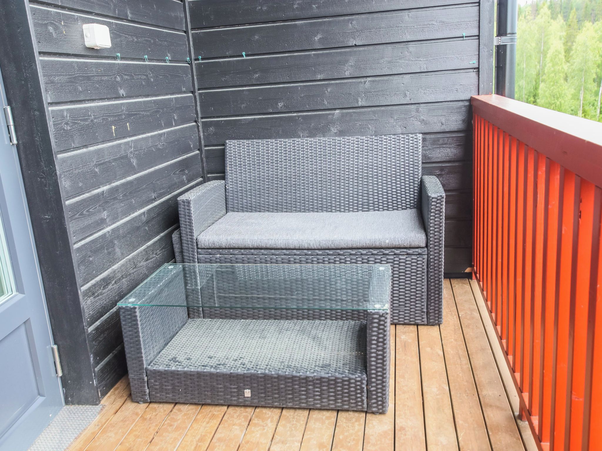 A wicker sofa with a glass table on a wooden deck with a red railing.
