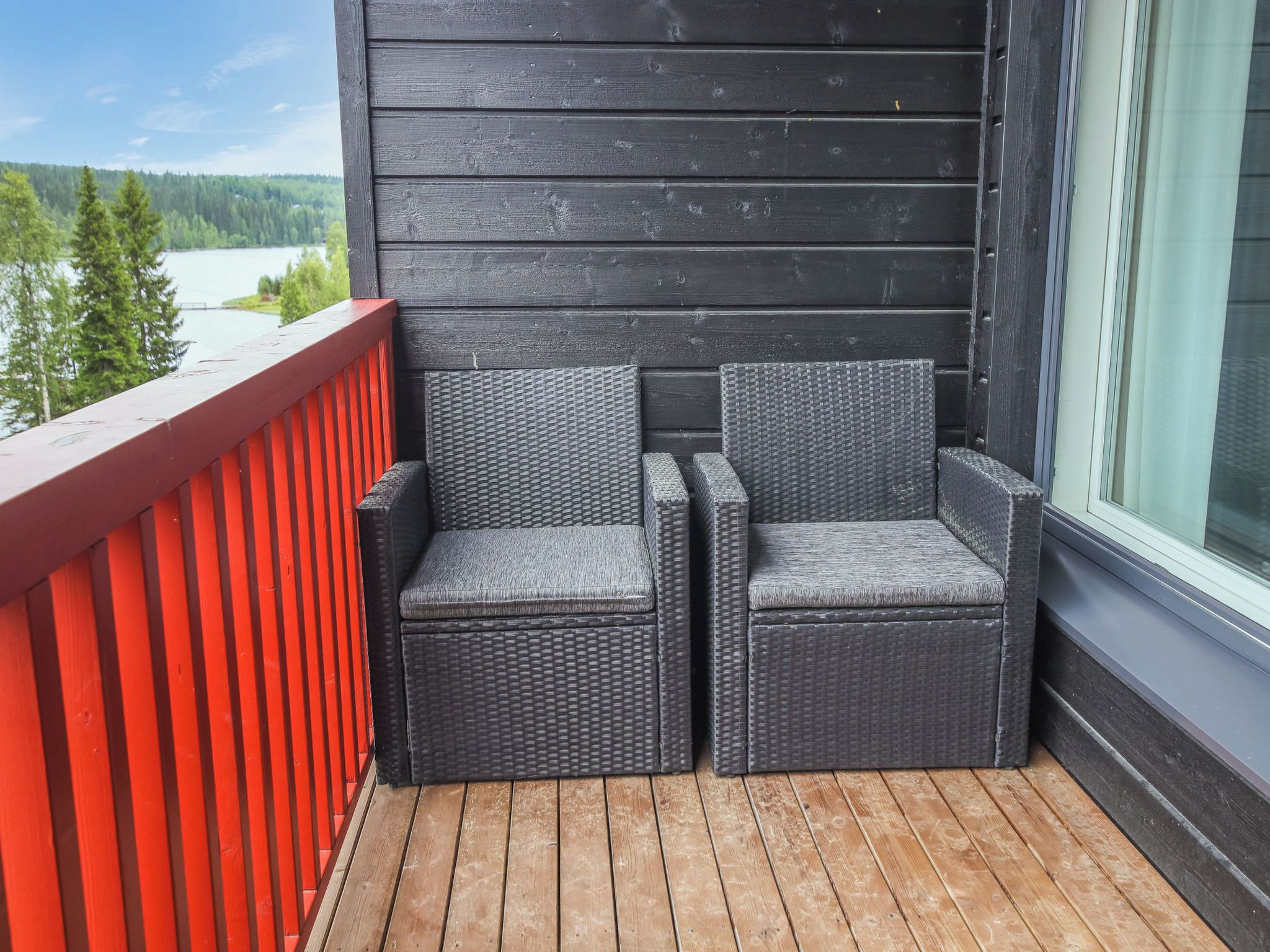 A balcony with two wicker chairs, a red railing, and a view of a lake and forest.