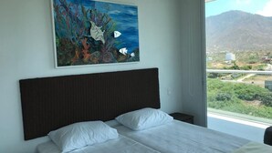 A bedroom with a bed, white bedding, a dark headboard, a painting of an underwater scene, and a view of a mountain and greenery through a window. - Apartamento Amoblado Playa Dormida Santa Marta (Santa Marta)