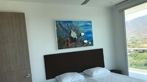 A bedroom with a bed, a nightstand, a painting of an underwater scene, and a view of a mountainous landscape through a window. - Apartamento Amoblado Playa Dormida Santa Marta (Santa Marta)