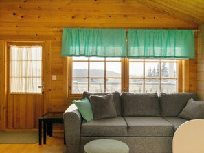 A cozy cabin interior with a wooden ceiling, a gray sofa with cushions, a small table, and a window with a view of a snowy landscape.