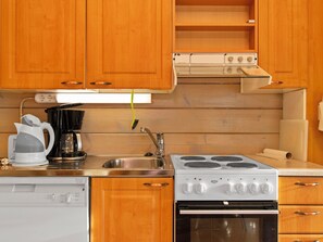A compact kitchen with wooden cabinets, a built-in oven, a stove, a sink, and a coffee maker. - Gasthalla a paritalo by Interhome (Hyrynsalmi)