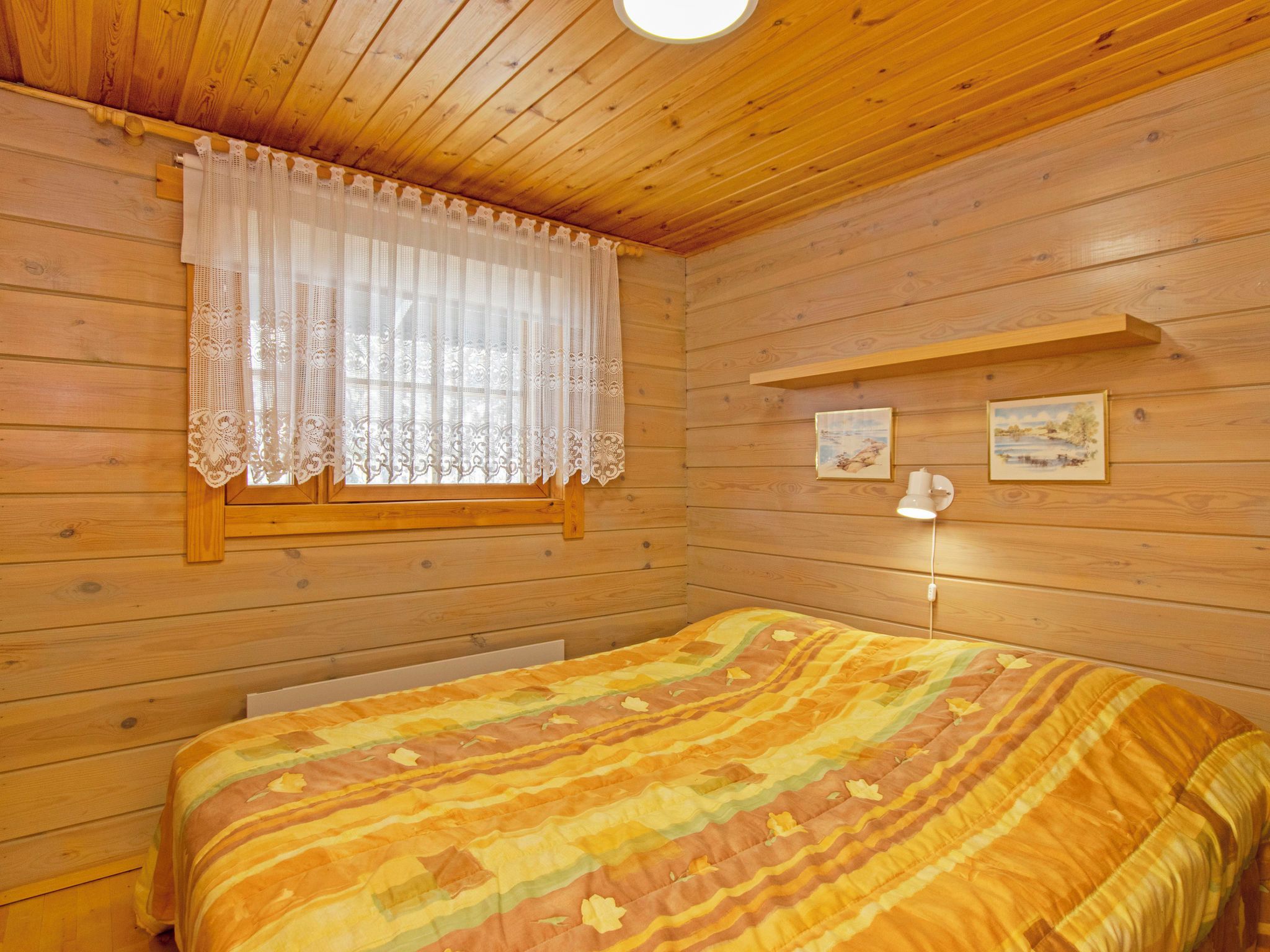 Una habitación de cabaña de madera con una cama, una ventana con cortinas de encaje y dos cuadros colgados en la pared.