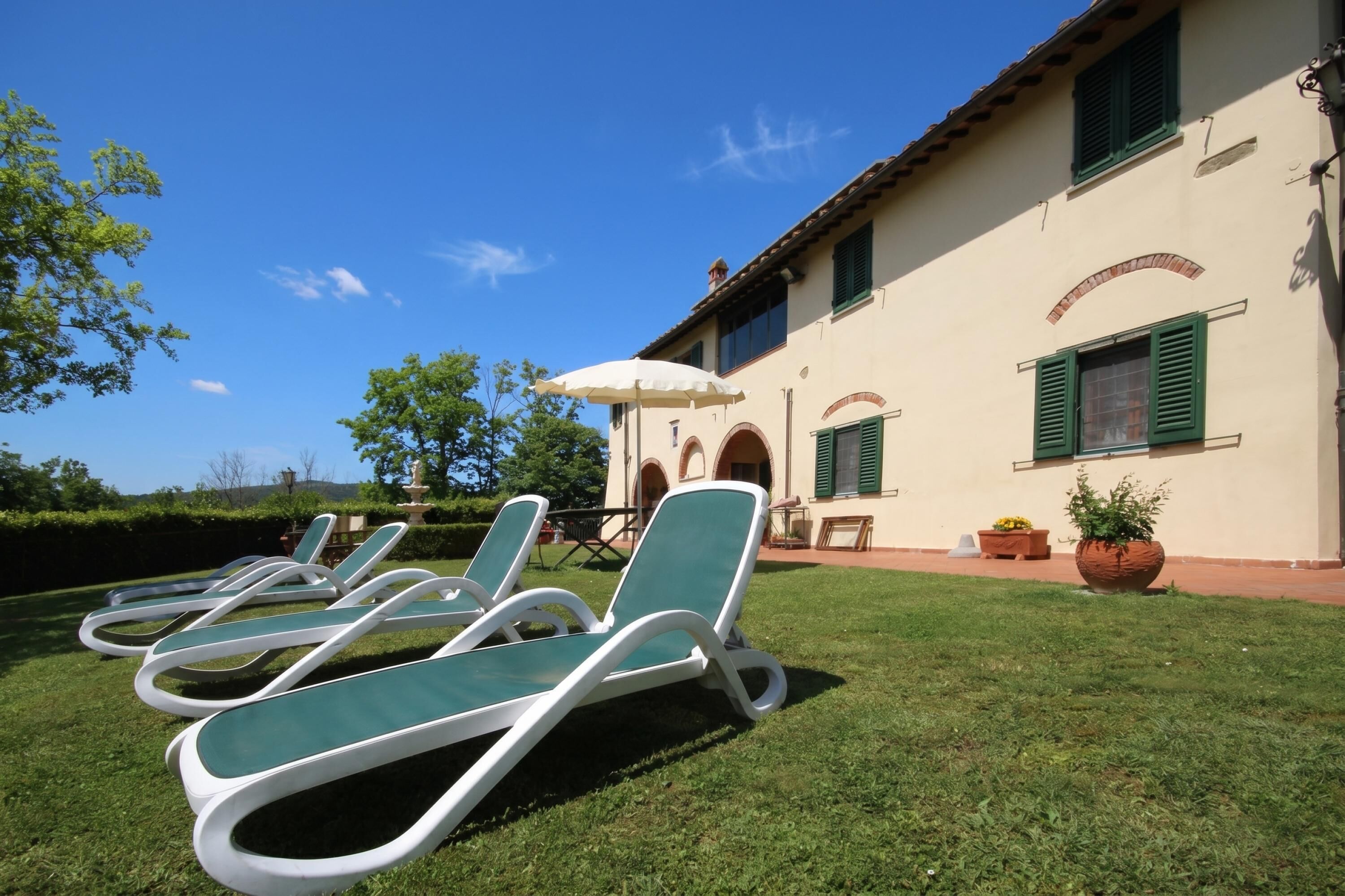 Eine Villa mit einer Terrassenanlage, die vier Lounge-Stühle, einen Tisch, einen großen Topfpflanzen und einen weißen Sonnenschirm zeigt.