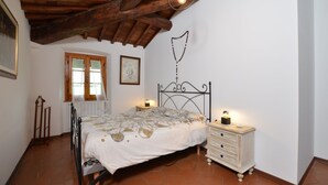 Une chambre avec un plafond en bois, un lit avec une tête de lit en métal, une table de chevet avec une lampe, une commode et une fenêtre avec des rideaux.