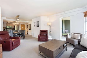 A modern living room with a sofa, armchair, and coffee table, a dining area with chairs, and a kitchen in the background.