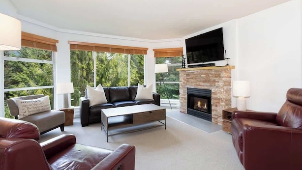 A modern living room with a fireplace, leather sofas, a flat-screen TV, and large windows. - Woodrun Lodge w/ pool & gym (Whistler)