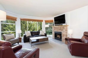 A modern living room with a fireplace, leather sofas, a flat-screen TV, and large windows.