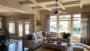 Ein großzügiges Wohnzimmer mit einer großen Eckcouch, einem Couchtisch und einem Deckenventilator. Große Fenster mit Vorhängen, ein Essbereich mit Tisch und Stühlen sowie eine Tür, die nach draußen führt.