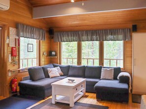 A cozy cabin interior with a sectional sofa, a white coffee table, and large windows offering a view of the forest. - Hyryn herra mökki by Interhome (Hyrynsalmi)