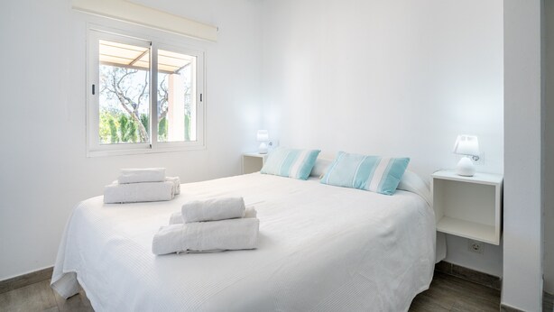 A bedroom with a large bed, two bedside tables with lamps, a window with a view, and neatly folded towels on the bed. - Vacation Home Ses Oliveres with Mountain View, Wi-Fi, A/C, Terraces & Garden (Pollença)