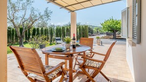 A covered patio with a table and chairs, a bottle of wine, and a view of the surrounding garden. - Vacation Home Ses Oliveres with Mountain View, Wi-Fi, A/C, Terraces & Garden (Pollença)