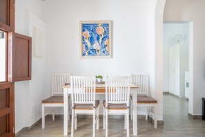 A dining area with a white table and chairs, a framed painting on the wall, and a kitchen in the background. - Vacation Home Ses Oliveres with Mountain View, Wi-Fi, A/C, Terraces & Garden (Pollença)