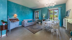 A room with teal walls, a white sofa, a dining table with chairs, a chandelier, and a window with curtains. - Holiday Apartment "La Punta Bellagio" with Lake View, Wi-Fi, Garden & Terrace (Bellagio)