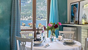 A dining table set with plates, cutlery, a bottle, and a vase with flowers, with a view of a mountainous landscape through a window. - Holiday Apartment "La Punta Bellagio" with Lake View, Wi-Fi, Garden & Terrace (Bellagio)