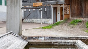 Un grifo con un estante metálico unido a un pilar de hormigón, un edificio de madera con base de piedra al fondo, y un letrero con texto.