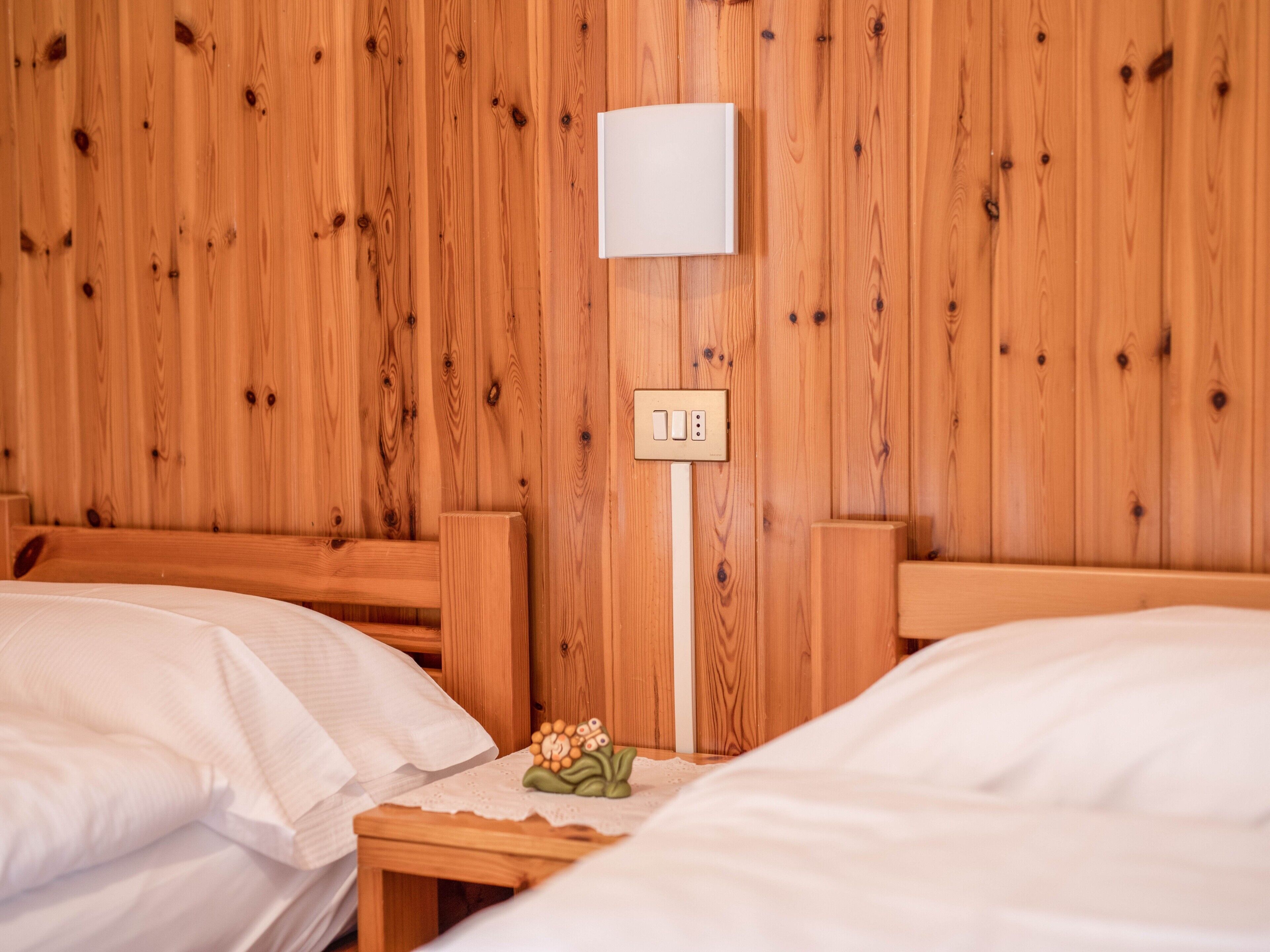 A wooden-paneled room with two beds, a nightstand with a lamp, and a wall-mounted light switch.