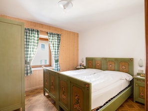 A bedroom with a wooden paneled wall, a bed with floral headboard, a nightstand, and a window with checkered curtains.