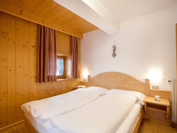 Un dormitorio con paneles de madera, una cama, una ventana con cortinas, una mesita de noche con lámpara y un aplique de pared.