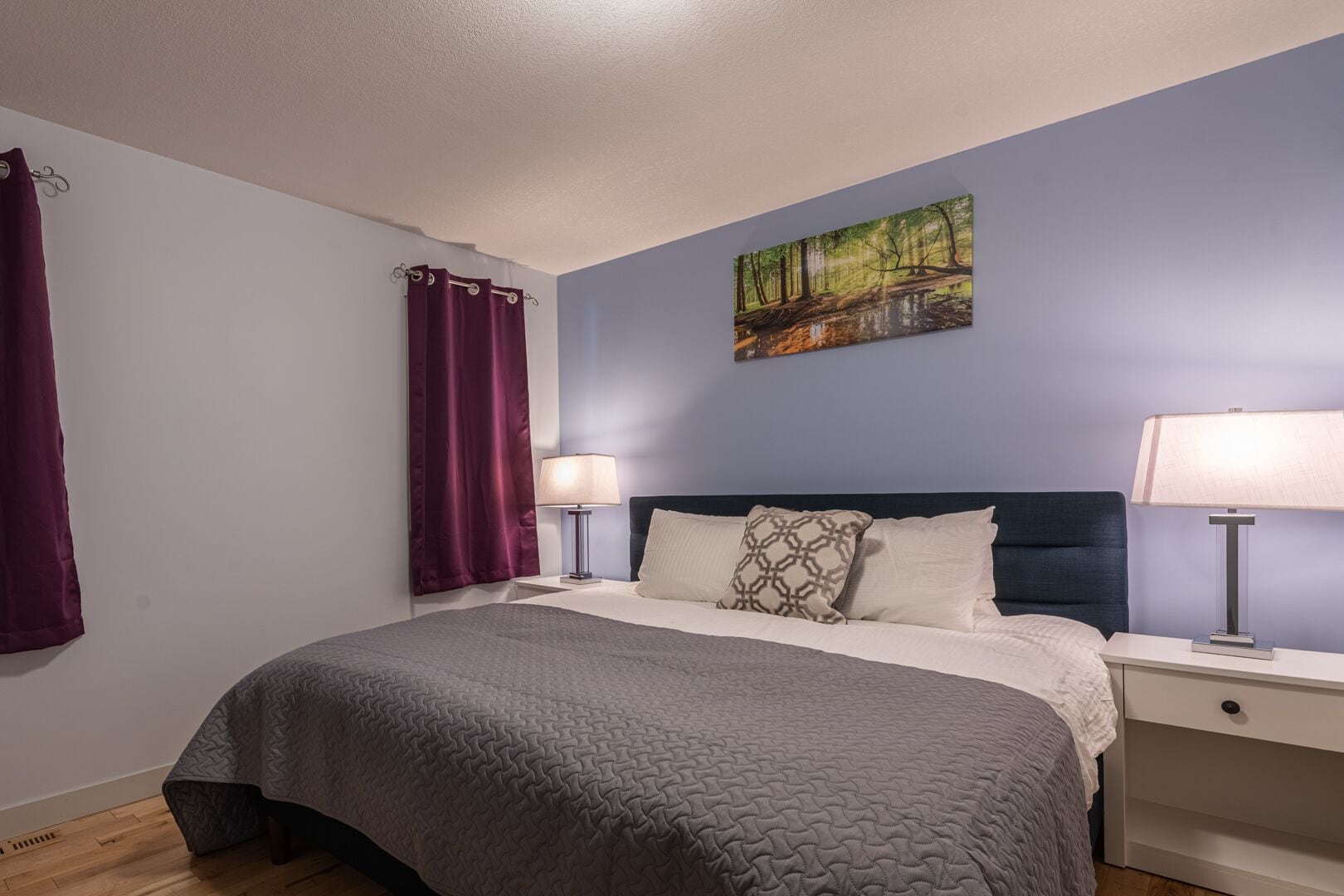 A neatly made bed with a grey quilt, two bedside lamps, a headboard, and a painting of a forest scene on the wall.