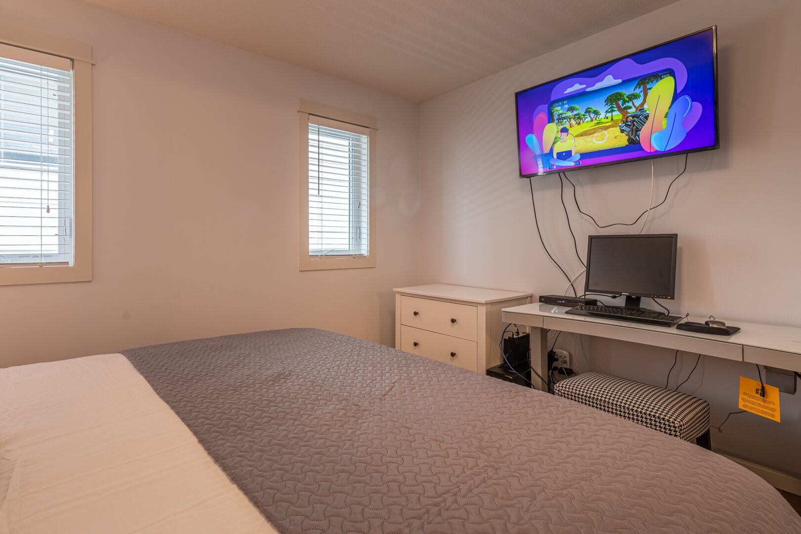 A bedroom with a bed, a desk with a computer, a television mounted on the wall, and two windows with blinds.