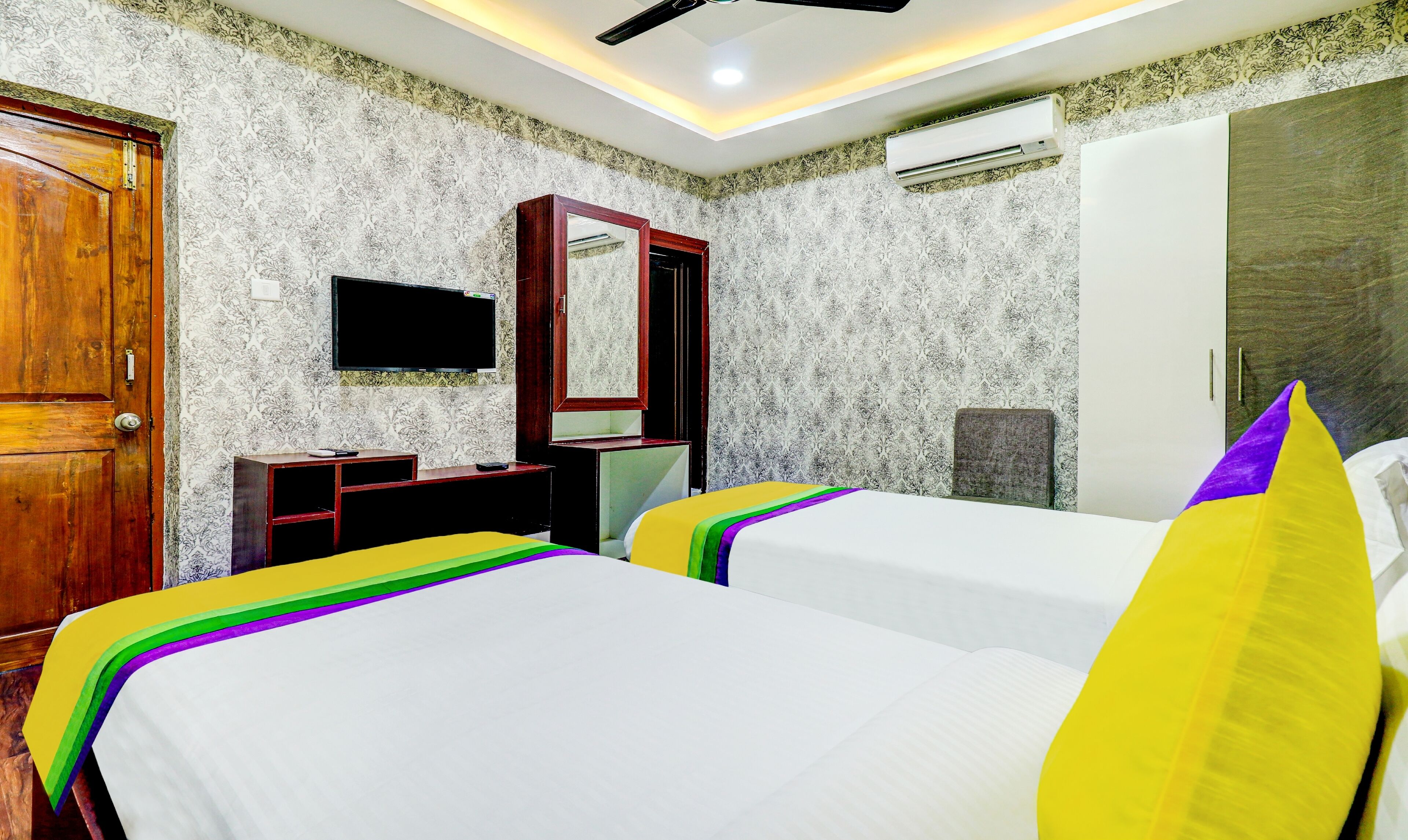 Habitación de hotel con dos camas, una puerta de madera, televisor, espejo y equipo de aire acondicionado.