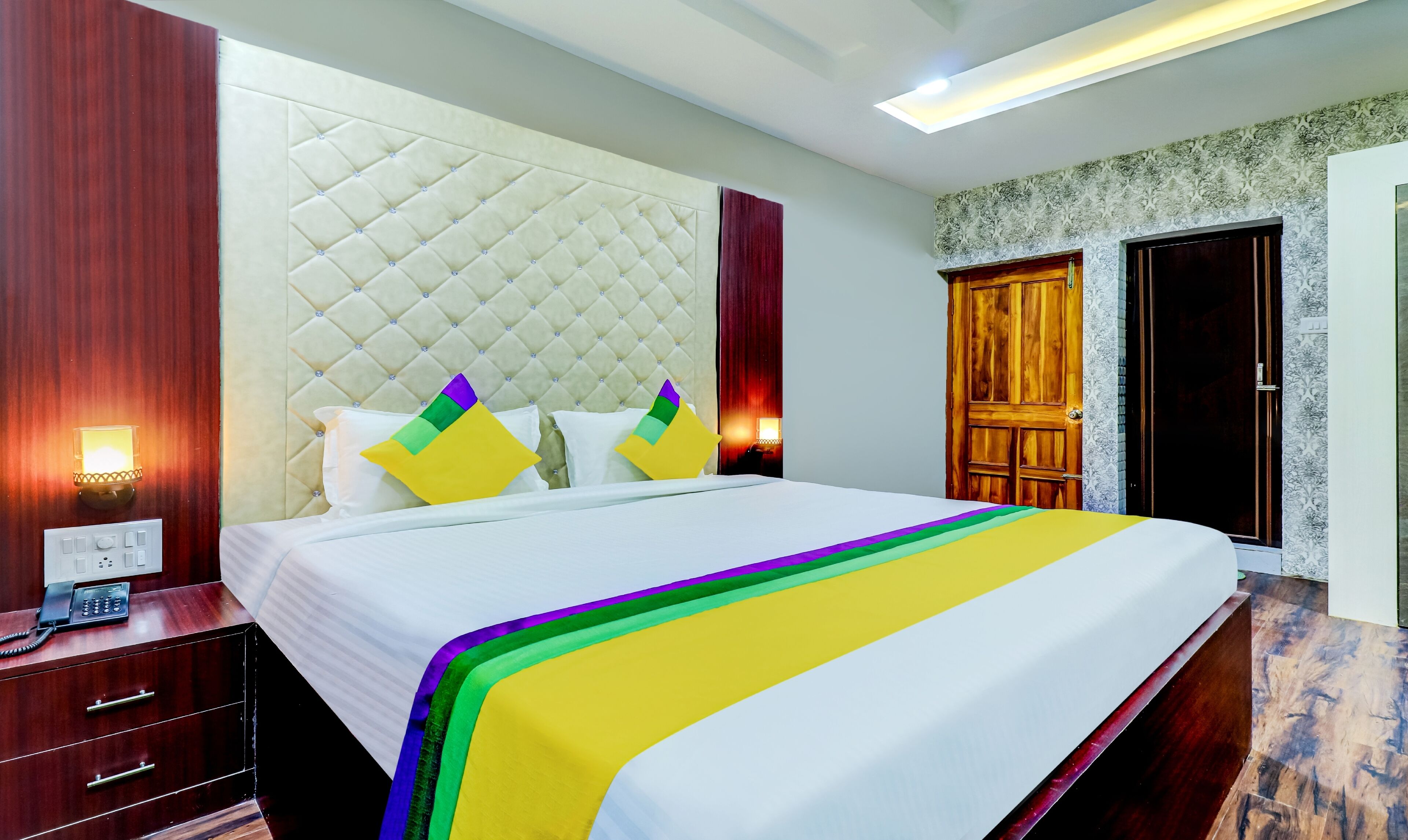 A hotel room with a large bed, a wooden wardrobe, a bedside table with a phone, and a wall-mounted light.