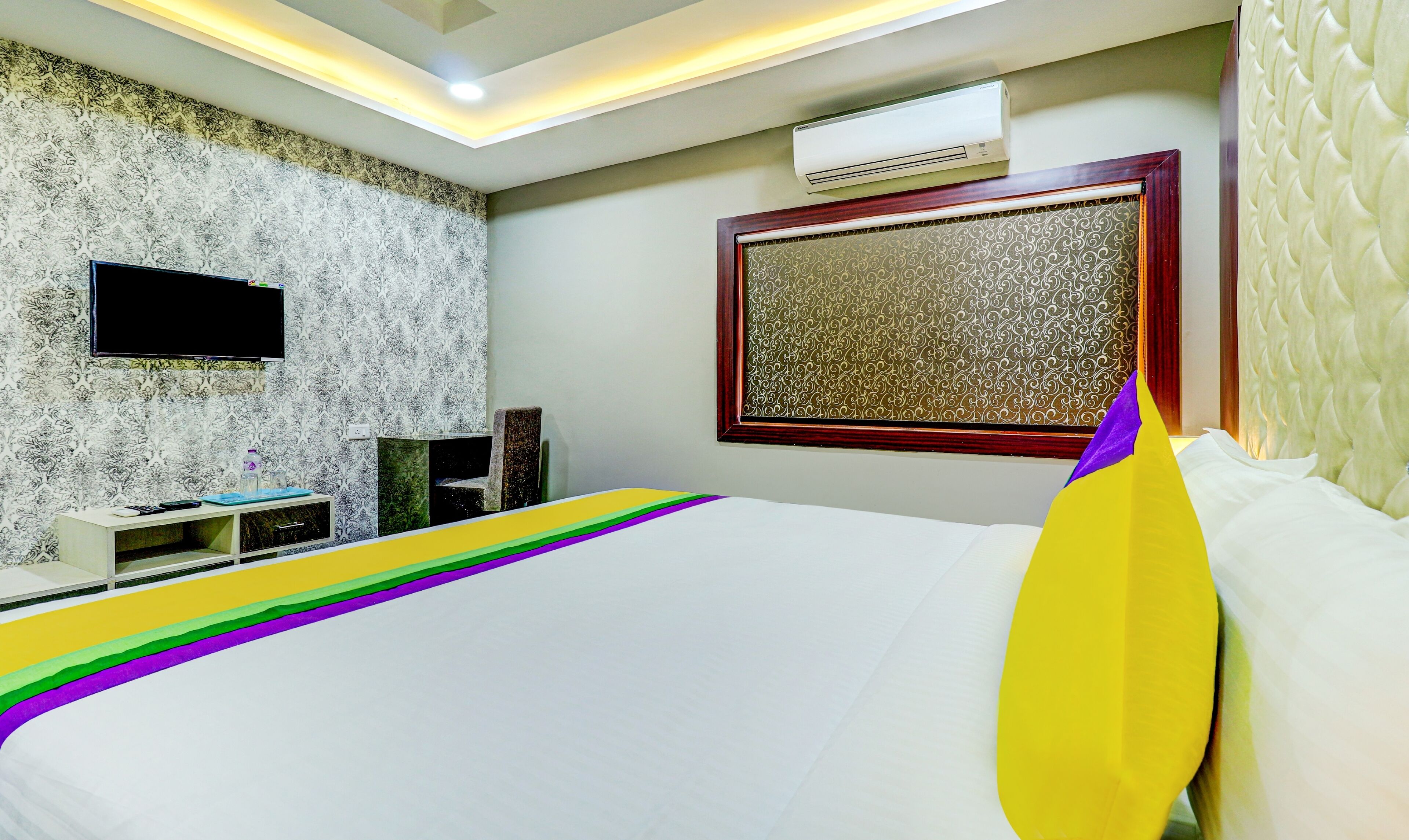 A hotel room with a bed, a TV, an air conditioner, and a patterned wall.