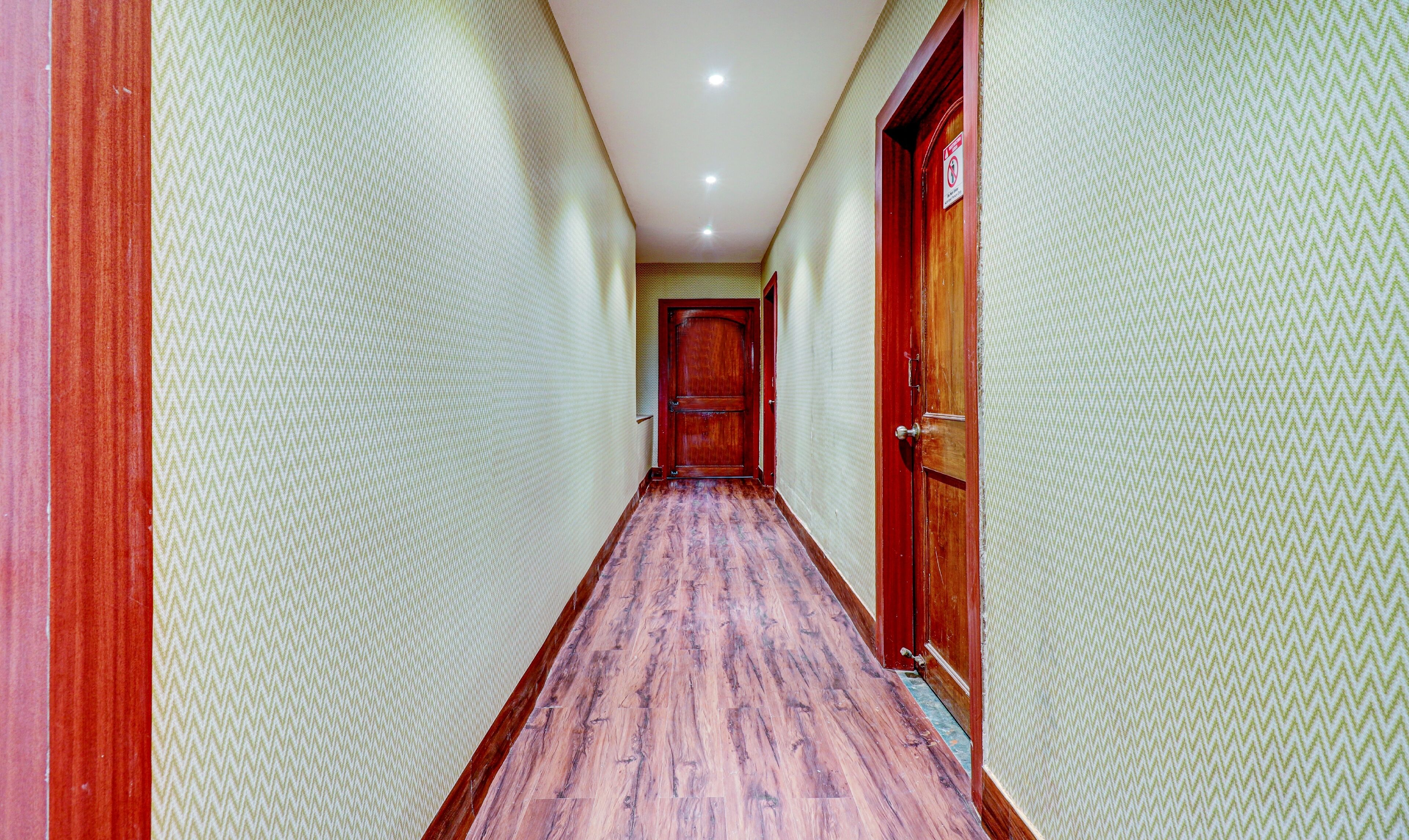 A narrow hallway with patterned walls and a wooden floor, leading to a door with a sign.