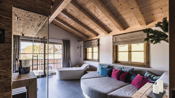 A modern interior with a wooden ceiling, a curved sofa with colorful cushions, a bathtub, and large windows.