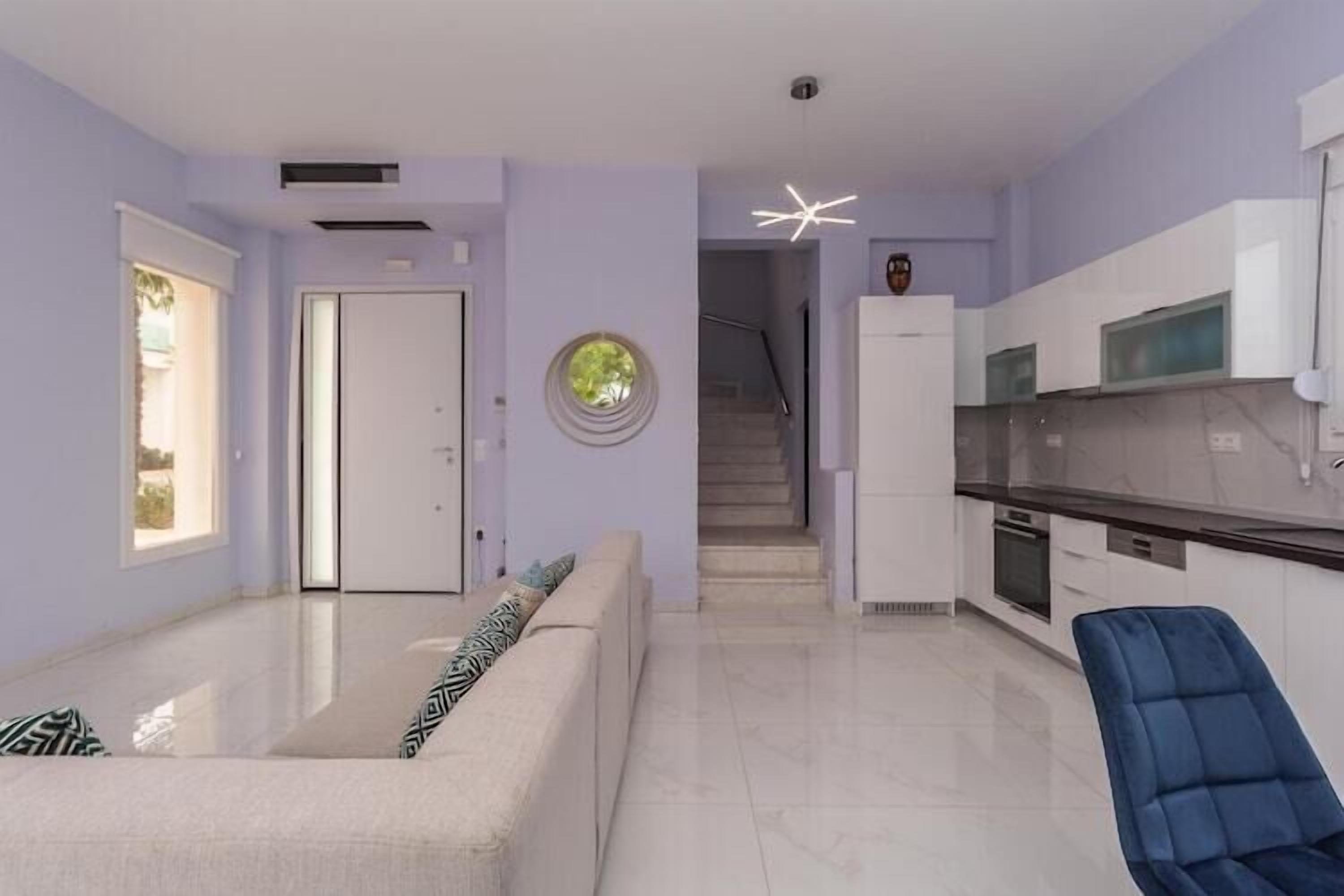 Un salon moderne avec un canapé, un fauteuil bleu, un coin cuisine doté de meubles blancs et un escalier menant à un autre niveau.