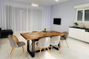 Un espace repas moderne avec une table en bois et des chaises recouvertes d’un tissu gris, une cuisine blanche avec une fenêtre, et une grande fenêtre en arrière-plan, ornée de rideaux légers et transparents.