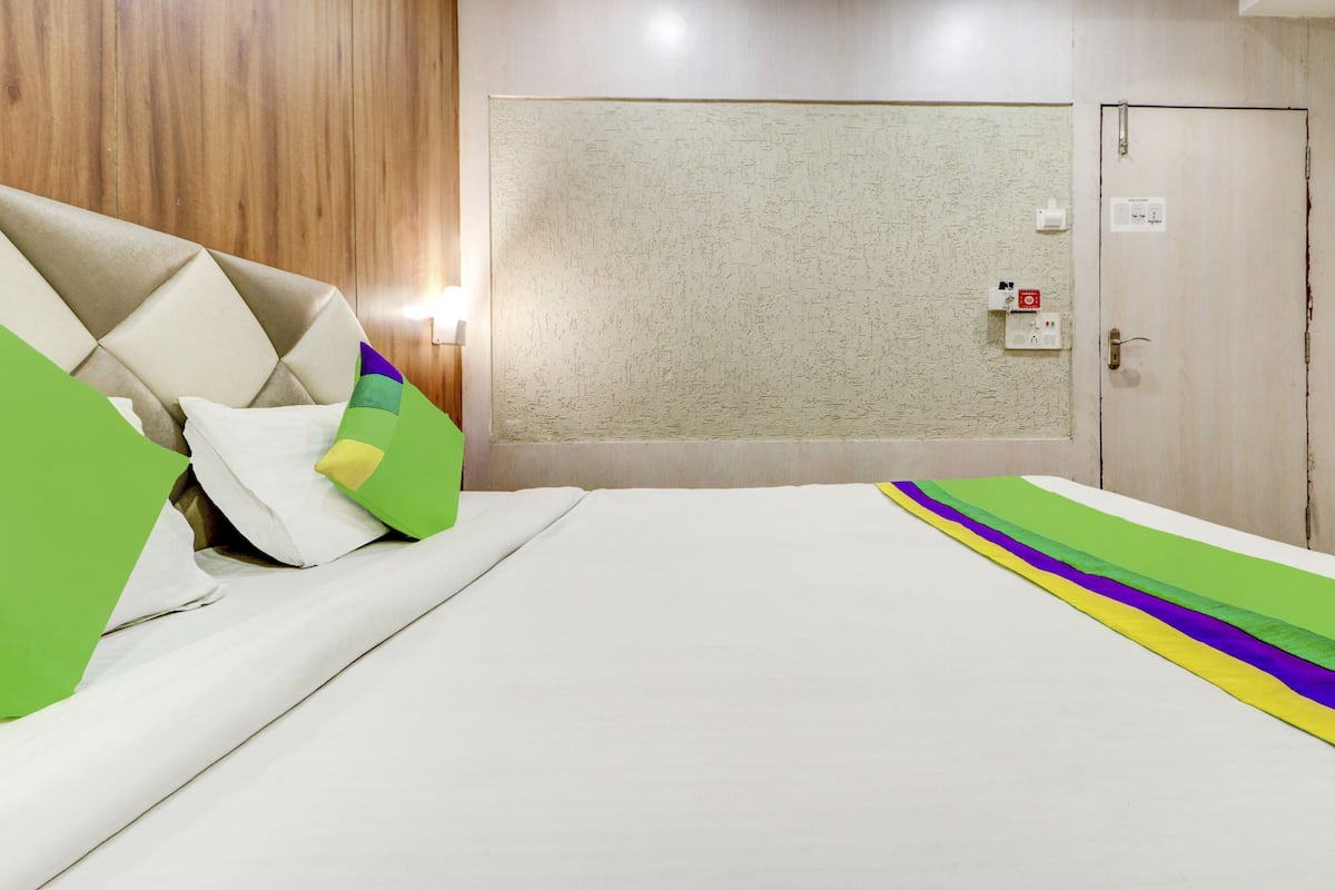 A hotel room with a bed, pillows, a wall-mounted light, a door, and a power outlet.