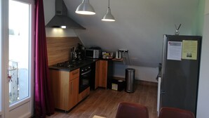 Une cuisine de taille réduite, dotée de meubles en bois, d’une cuisinière, d’un four à micro-ondes et d’un réfrigérateur. On y trouve deux lampes suspendues, une fenêtre avec des rideaux et une poubelle.