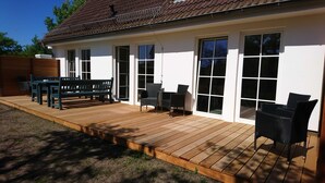 Una casa con terraza de madera, zona de comedor con sillas y mesa, y un porche cubierto con más asientos.