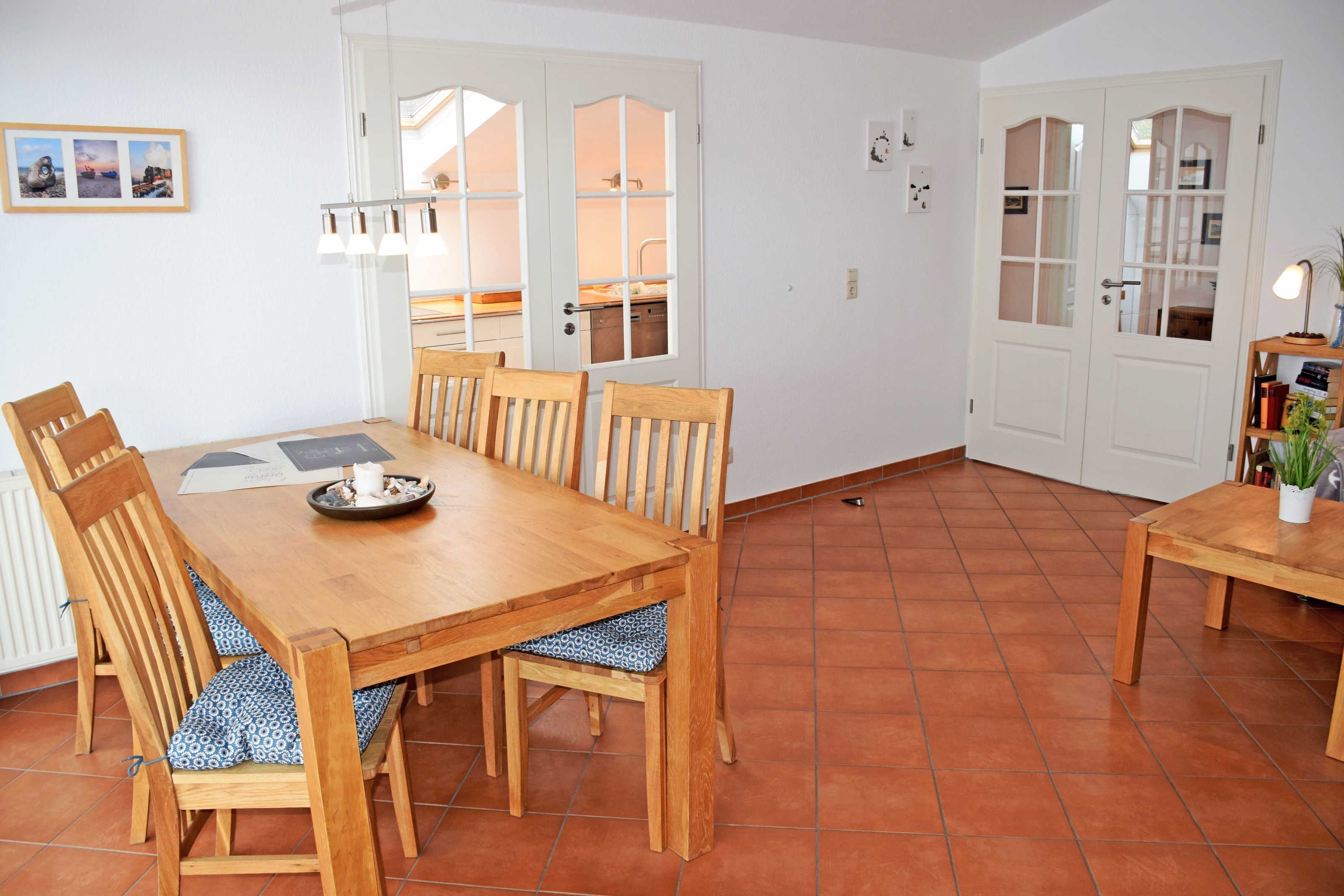 A dining area with a wooden table and chairs, a tiled floor, and a view of the kitchen through a glass door.