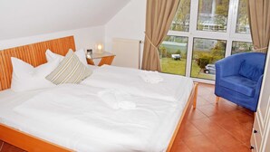 A bedroom with a large bed, a nightstand, a blue chair, and a view of a garden through a window. - Fewo mit Balkon und Strandkorb am Strand (Baabe)
