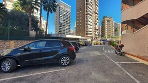 A parking area with cars and a motorcycle, surrounded by tall buildings and a fence with palm trees. - LAS ADELFAS (2 BEDROOM) (Benidorm)