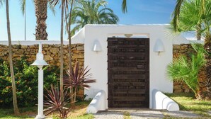 Un edificio blanco con un muro de piedra y una puerta de madera. Hay dos lámparas blancas a cada lado de la puerta.
