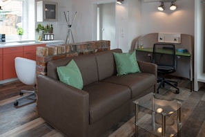 Un salon moderne avec un canapé en cuir marron, des coussins verts, une table basse en verre, une cheminée en briques, un coin cuisine doté d’un plan de travail blanc et un fauteuil de bureau noir.