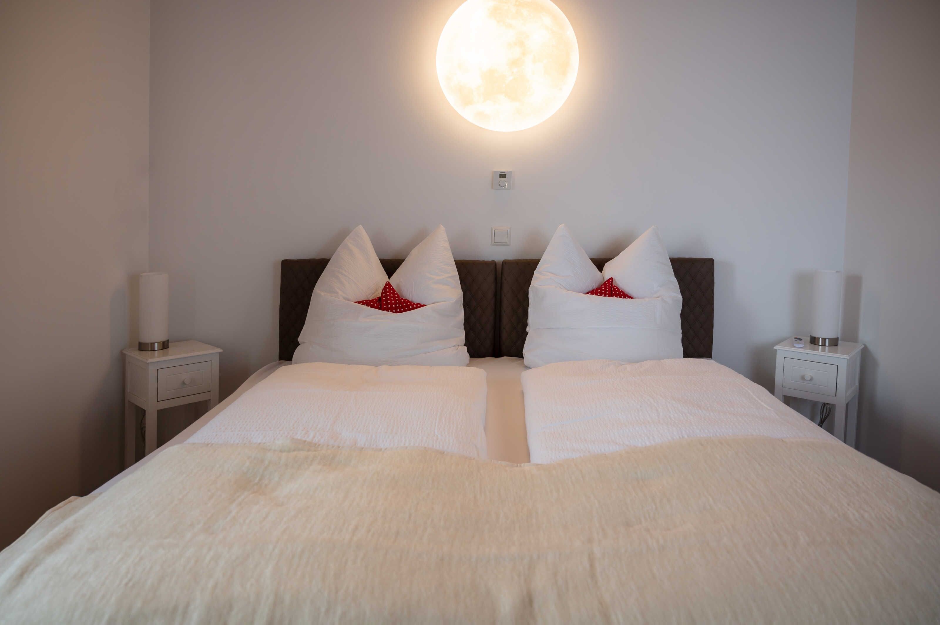 A neatly made bed with white linens and red pillows, a wall-mounted round light, and two bedside tables with lamps.