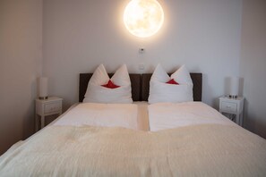 Een net opgemaakt bed met witte lakens en rode kussens, een rond wandlamp en twee nachtkastjes met lampen.