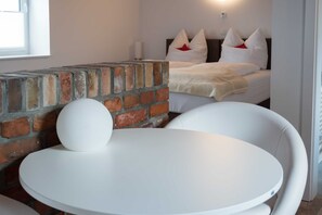 Une pièce avec un mur en briques, une table ronde, un lit avec une literie blanche et des oreillers rouges, et une chaise blanche.