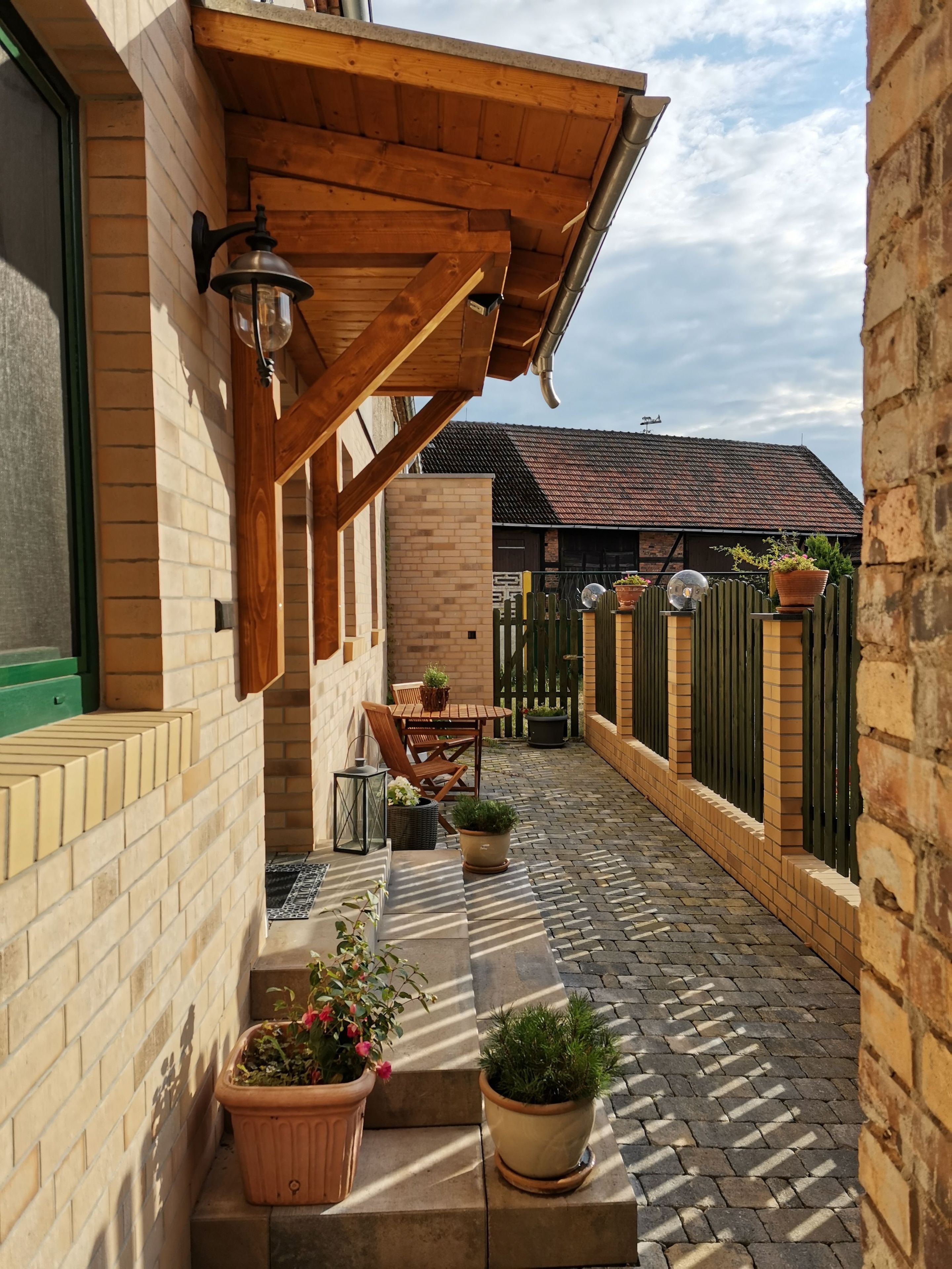 A covered patio with a brick wall, wooden pergola, outdoor seating, and potted plants.